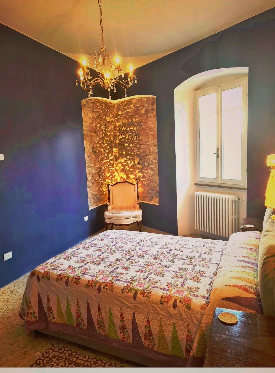Second bedroom at Corte Averaldo — deep blue walls, gold crystal chandelier, 16th century stone wall feature, arched window with natural light, San Felice del Benaco Lake Garda Italy